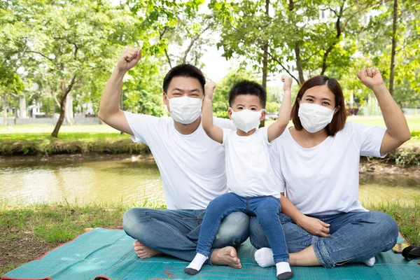 Asian family wearing protective medical mask for prevent virus Covid-19 and hand up and sitting together on floor at green public garden. Family protection from contaminated air concept