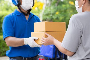Yüz maskesi takan Asyalı kurye ve motosikletli mavi üniformalı kadın müşteriye paket kutusu servisi yapıyor.