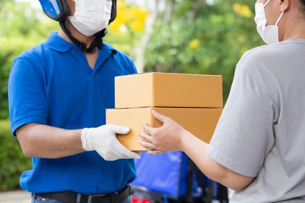 Asian delivery man wearing face mask and blue uniform with motorcycle delivering parcel box express service to woman customer