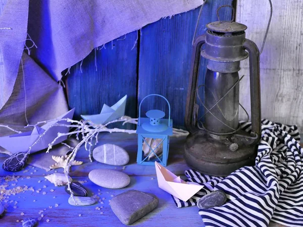 Installation of decorative lantern, sea stones, seashells, paper boats, sea salt on a wooden table, textural background, concept of sea travel, blue lighting, top view