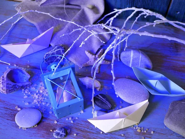 Installation of decorative lantern, sea stones, seashells, paper boats, sea salt on a wooden table, textural background, concept of sea travel, blue lighting, top view