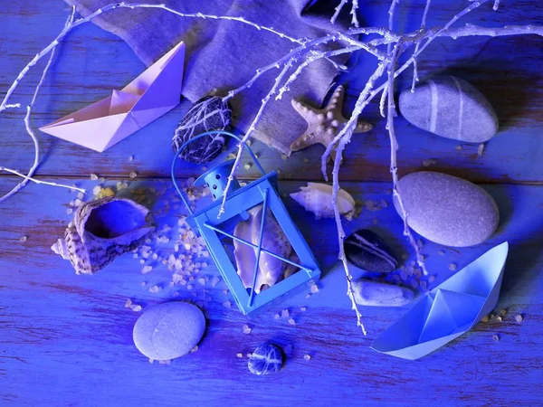 Installation of decorative lantern, sea stones, seashells, paper boats, sea salt on a wooden table, textural background, concept of sea travel, blue lighting, top view