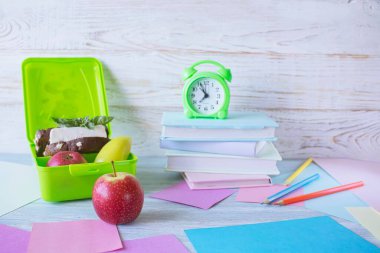 Okul konseptine geri dönelim, okul yemekleri. Sağlıklı sandviçle beslenme çantası, çalar saat, beyaz ahşap arka planda bir yığın kitap ve elma..