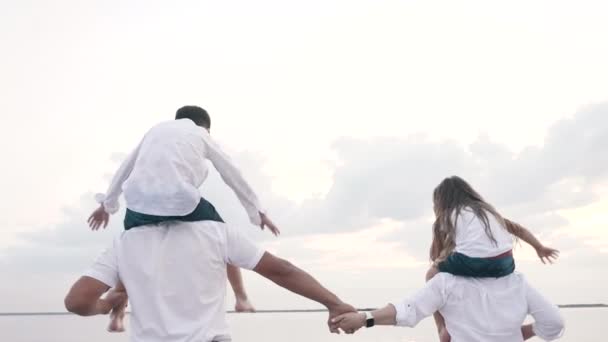 Glückliche Familie mit Kindern auf Schultern, die Händchen halten, gemeinsam im Freien spazieren gehen — Stockvideo