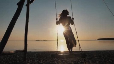 Genç kaygısız güzel kız salıncakta sallanıyor, cennet denizinin tadını çıkarıyor gün batımında rahatlıyor