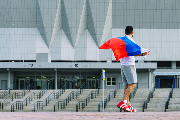A young guy with a beard in his white T-shirt and shorts is holding a flag of Russia that is developing in the wind