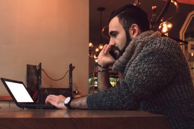 Bir sakallı bir genç adam beyaz bir perde arkasında bir dizüstü bilgisayarda bir kafede bir masada çalışıyor