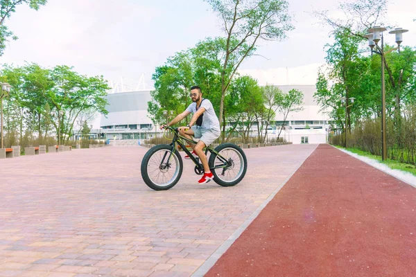 Yaz aylarında sıcak, güneşli bir günde şehir parkı ile sakal ve güvenle büyük bir bisiklet üzerinde güneş gözlüğü takmış genç bir adamla skates