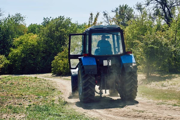 在炎热的夏日里 一辆带大轮子的蓝色拖拉机在森林中央的一条尘土飞扬的泥土路上骑着 — 图库照片