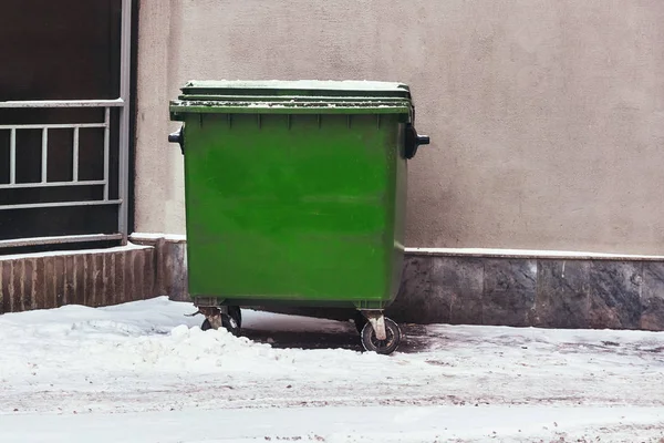 Green garbage can of plastic in winter in the snow near the wall in the city