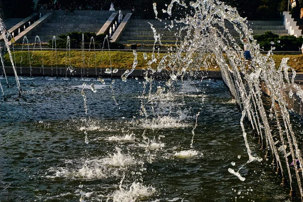 Water jets of a large fountain shine in the rays in the central city park of the city in the air on a sunny summer day — Stock Photo, Image