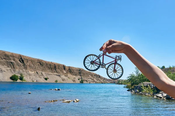 Czerwony zabawka metalowa rower w rękach na tle klifu w oddali z złudzenie optyczne duży rower na jasny słoneczny dzień w lecie podczas podróży — Zdjęcie stockowe