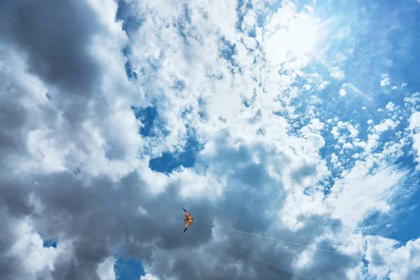 Um papagaio grande flutua alto no céu entre as nuvens contra o céu azul e o sol de verão brilhante e é controlado por duas cordas brancas que vão para o chão — Fotografia de Stock