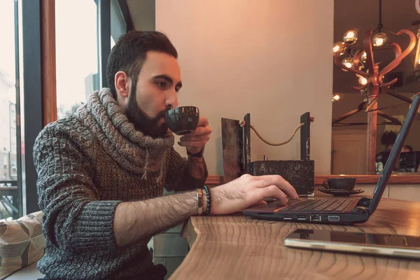 Молодой парень с бородой и в теплом свитере сидит в уютном кафе и работает на ноутбуке в Интернете — стоковое фото