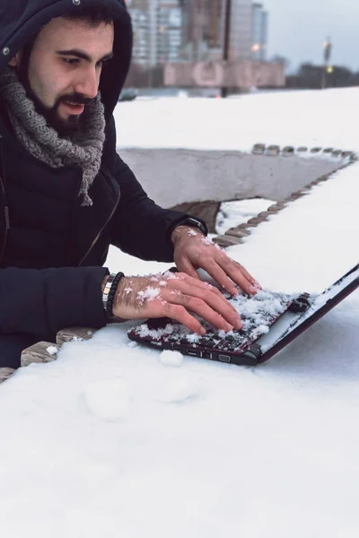 Jovem com uma barba em uma jaqueta quente trabalha com um laptop ao ar livre na neve em um dia gelado de inverno Imagem De Stock