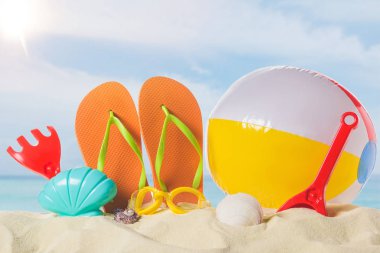 Flip flops with beach ball and toys in sand on blue sky background clipart