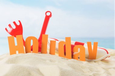 Beach toys and Holiday inscription in sand on blue sky background clipart