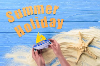 Woman holding toy boat by Summer holiday inscription on blue wooden background clipart