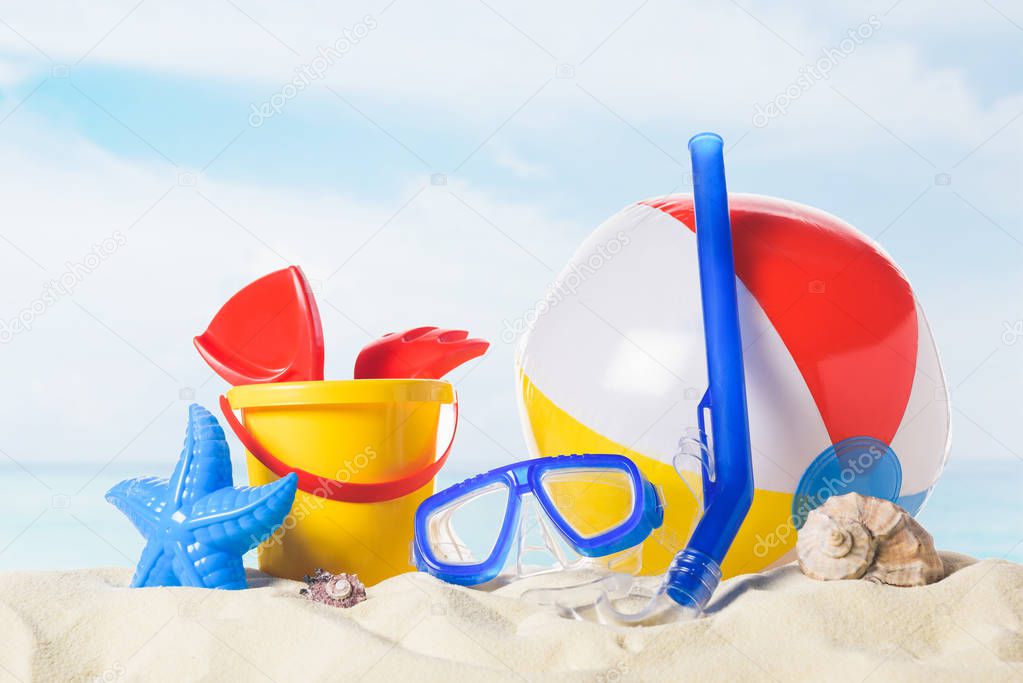 Diving mask with beach ball and toys in sand on blue sky background