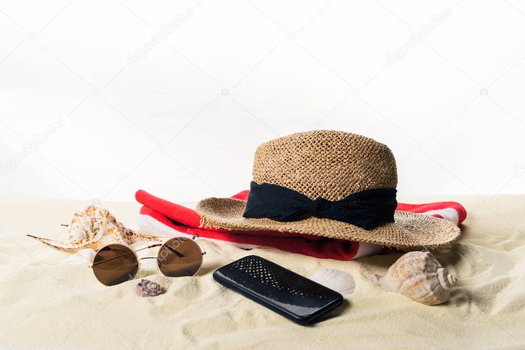 Straw hat on towel and sunglasses with seashells in sand isolated on white
