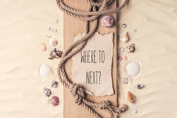 Summer travel template with seashells and rope on wooden pier on light sand with "where to next?" lettering