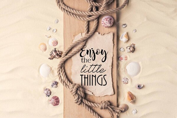 Summer travel template with seashells and rope on wooden pier on light sand with "enjoy the little things" inspection