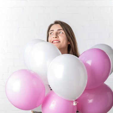 Pretty girl smiling over bunch of balloons on white brick wall background clipart
