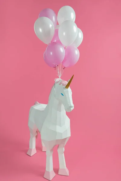 Toy unicorn with tied bunch of balloons on pink background