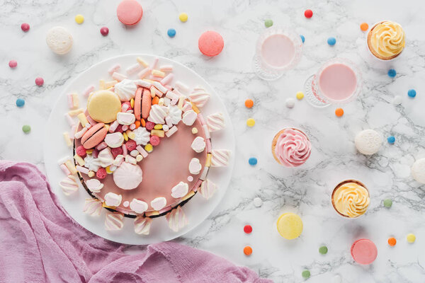 top view of pink birthday cake with marshmallows, cupcakes and milkshakes on marble table 