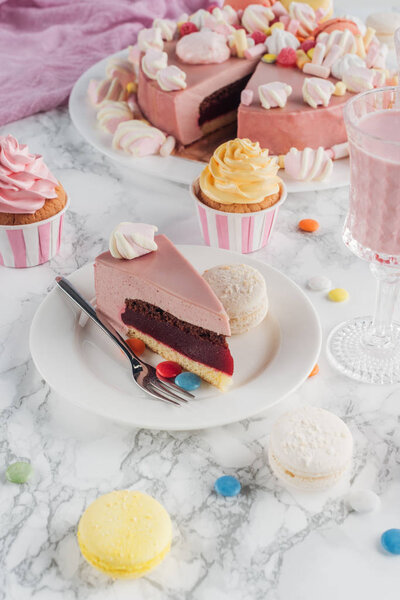 piece of birthday cake, macaroons, colorful cupcakes and milkshake in glass on table 