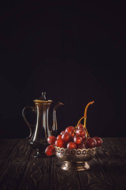 close-up shot of red grapes in vintage metal bowl and teapot on wooden table on black clipart