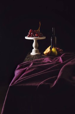 still life with fruits and bottle of wine on burgundy drapery on black clipart