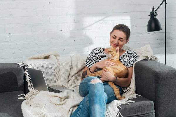 beautiful smiling young woman sitting on sofa and holding cute red cat