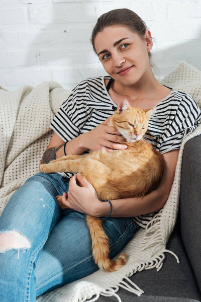 красивая улыбающаяся молодая женщина держит красную кошку и сидит на диване
