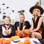 Porträt von Mutter und Kindern in Halloween-Kostümen mit Kürbissen in den Händen, die zu Hause auf dem Sofa sitzen