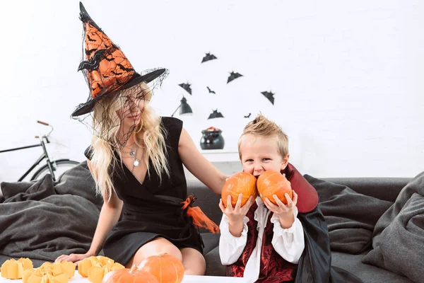 portrait of little boy in vampire halloween costume biting pumpkins sitting on sofa with mother in witch costume