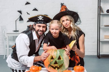 portrait of parents and little daughter in halloween costumes at table with pumpkins at home clipart