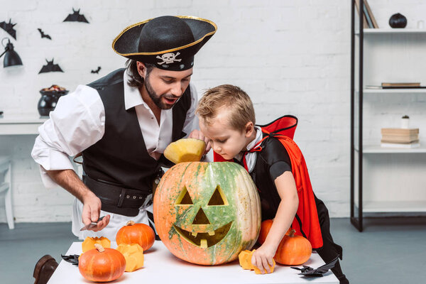 мужчина в пиратском костюме и сын в костюме вампира на Хэллоуин, смотрят в тыкву вместе дома
