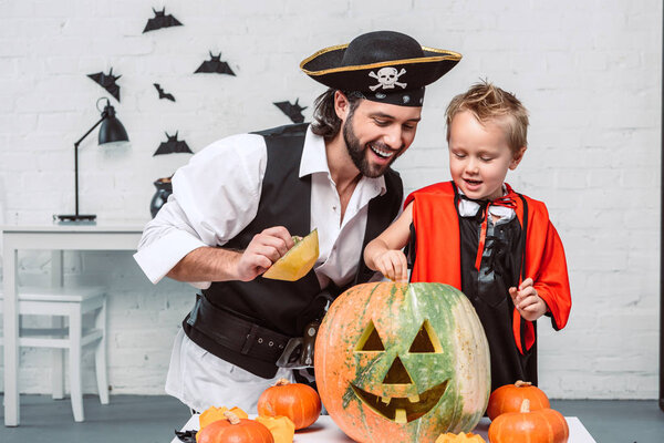 Улыбающийся мужчина в костюме пирата и сын в костюме вампира на Хэллоуин, смотрящий в тыкву вместе дома
