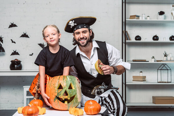 портрет улыбающихся отца и дочери в костюмах на Хэллоуин за столом с тыквой дома
