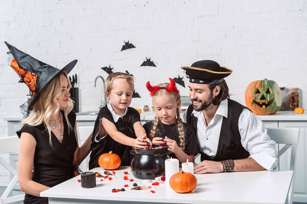 family in various halloween costumes at table with black pot with treats in kitchen at home