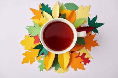 top view of cup of tea and colorful handcrafted leaves on white surface clipart