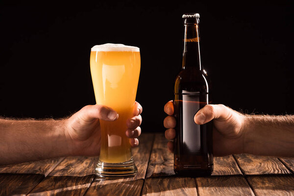 cropped image of two men clinking by glass and bottle with beer at wooden table 