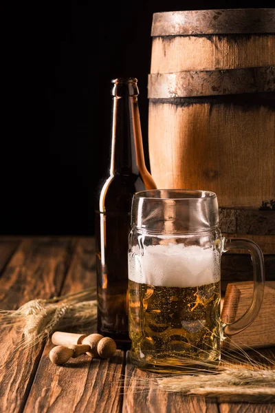 mug of fresh beer with foam, beer bottle, wheat and wooden barrel at table on black background