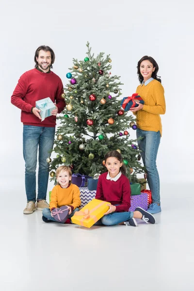 Happy parents with gift boxes standing while siblings sitting near christmas tree isolated on white