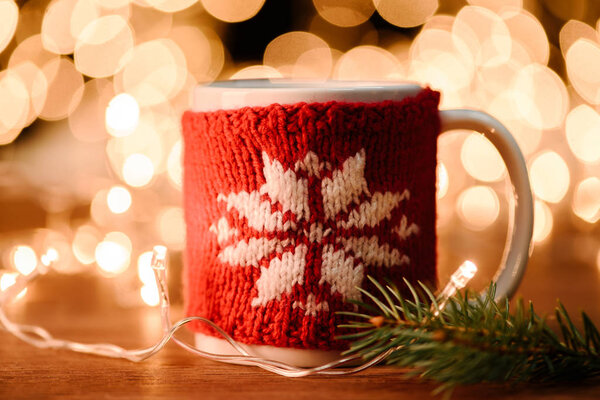 close up view of cup of hot drink, pine tree branch and garland on wooden tabletop with bokeh lights background