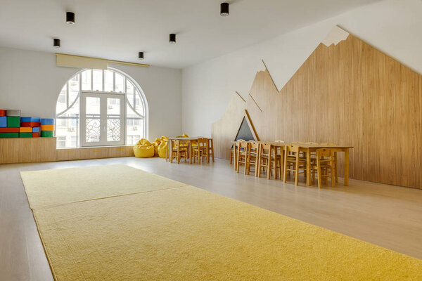 интерьер игровой комнаты с деревянными стульями и столами, желтый ковер, кресла из бобов и светлые блоки в детском саду
