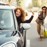 Kızıl saçlı kıvırcık kadın uzak ve sallanan el ile City tekerlekli çanta ile şık kadın turist sokak sürüş