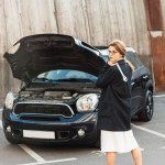 Female woman in coat and eyeglasses posing with spanner near car with opened hood at urban street