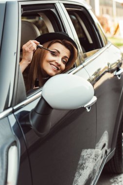 happy adult woman in black hat looking at wing mirror while doing make up in car